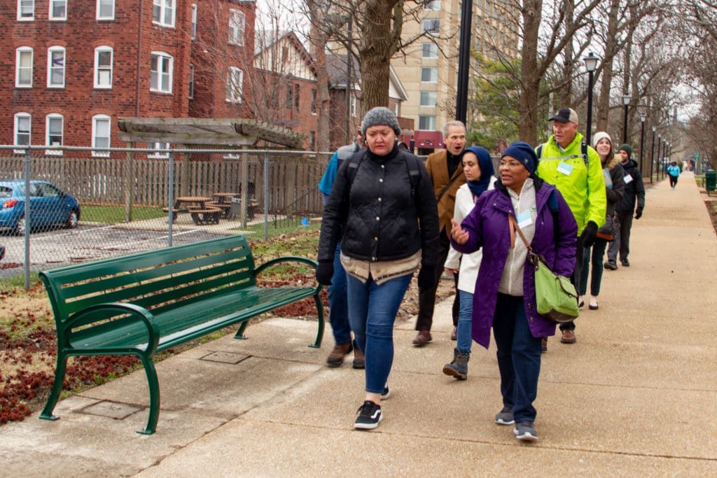 A diverse group of people wearing winter clothes walking along a path next to a green bench in a residential neighborhood. 
