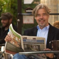 A medium shot photo of Jay Walljasper sitting outside a cafe on the sidewalk reading a newspaper. 