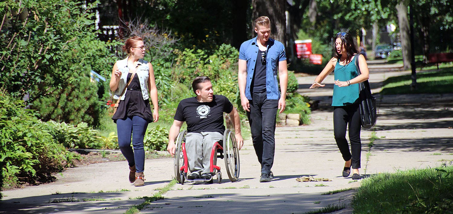 Four young people moving and walking on the sidewalk, one person uses a wheelchair. 