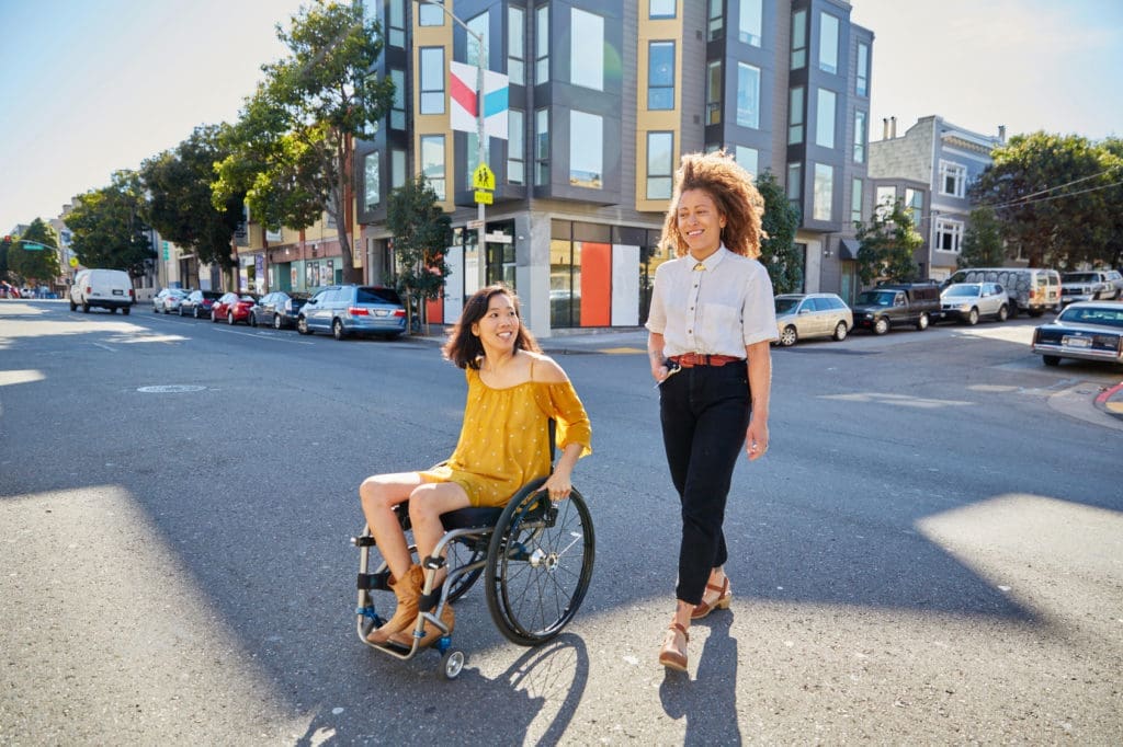 Two younger adults rolling and walking in the middle of an urban setting with a modern high rise building in the background. One person is using a wheelchair. 