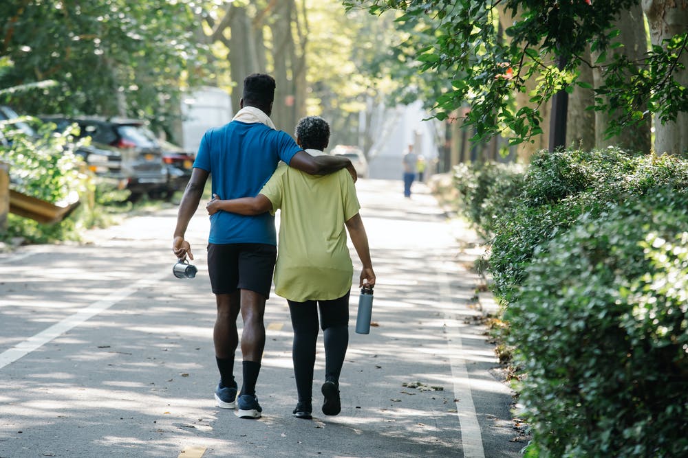 A young Black man and an older Black woman walk away from the camera on a tree-lined street. They appear to be son and mother and are out on a walk together. 