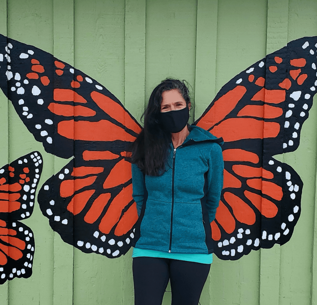 Janet Castanos wears a mask and stands in front of a mural of monarch butterfly wings.