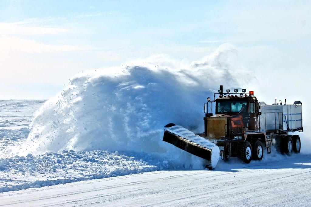 A huge snow plow is shown blasting snow all over the sides of a road. 