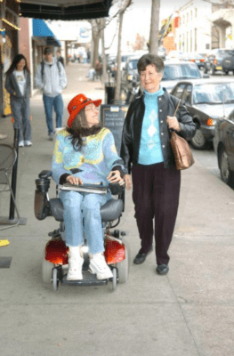 Two older women — one walking, one using a wheelchair — on a city street.