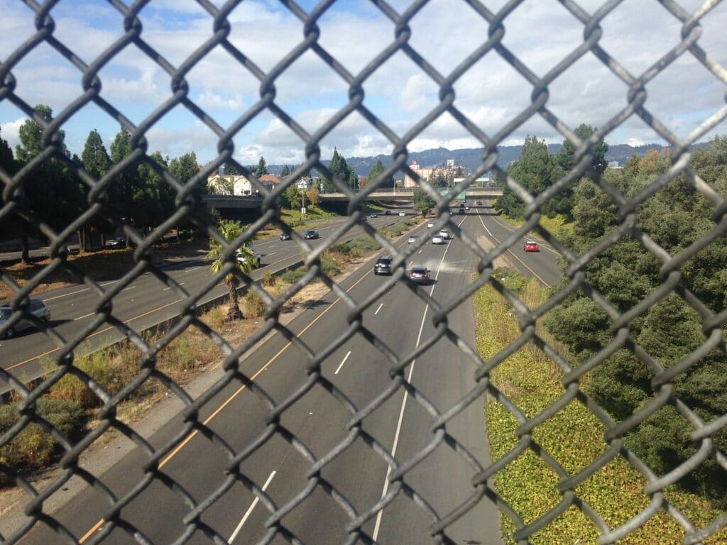 The viewer overlooks a controlled access highway through a chain link fence on a bridge. The reconnecting communities program was designed to begin to remove these dividers.