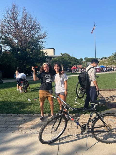 Kenedy and Hosea stand smiling with a bicycle on a sunny campus lawn during their outdoor event.