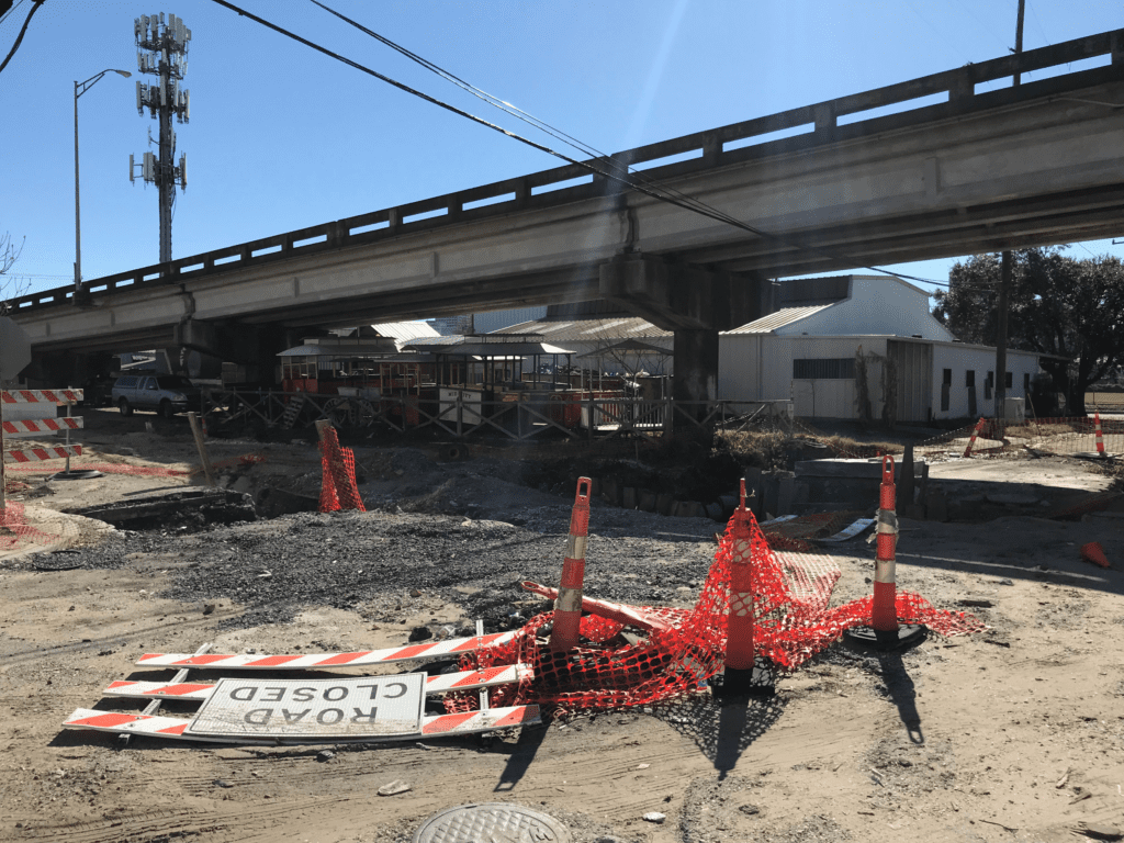 A road closed barricade lies in the dirt alongside cones and plastic barrier fence. A concrete overpass is above the construction.