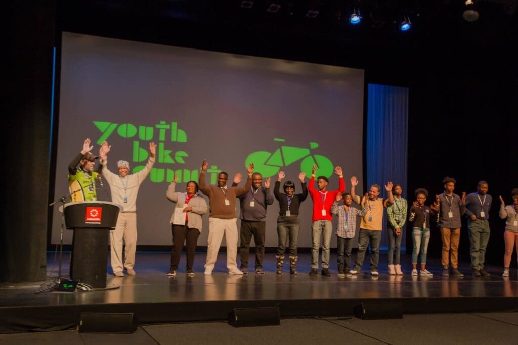 A group of young people on a stage are holding their hands up. The background is a screen of the Youth Bike Summit.