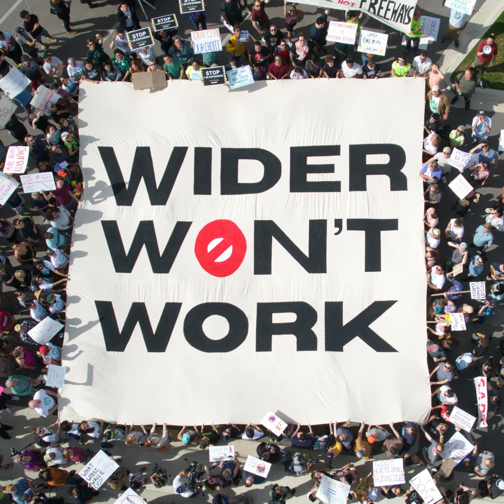 advocates hold a banner overhead for a drone photo saying "wider won't work"