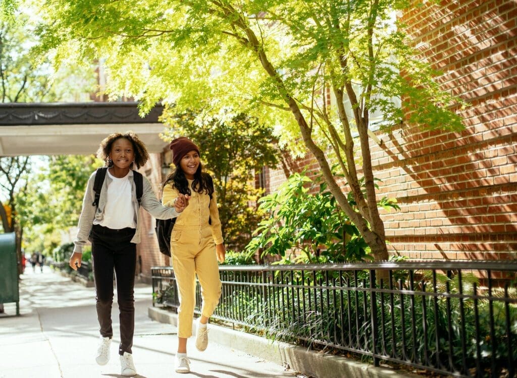 two young girls walk side by side holding hands down a nice tree-shaded street