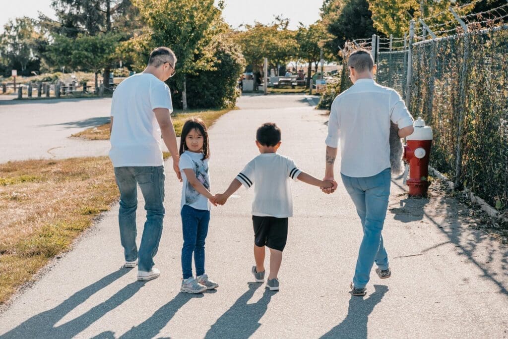 on a sidewalk in an urban area, two adults hold the hands of two children, one child looks back at the camera