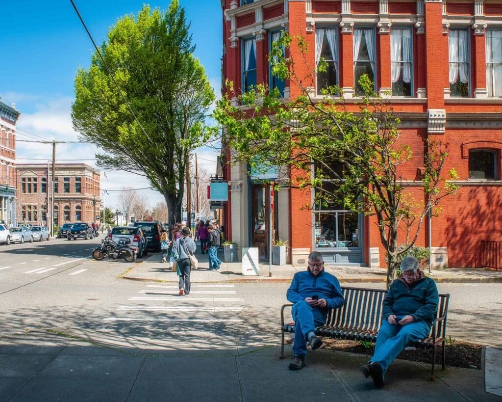 Two people sit on a bench along a street while another person crosses a street in a crosswalk behind them in Port Townsend, Washington. The sidewalk is bustling with people.