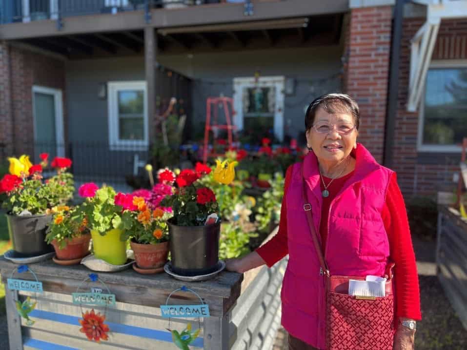 a woman stands alongside potted flowers behind an apartment