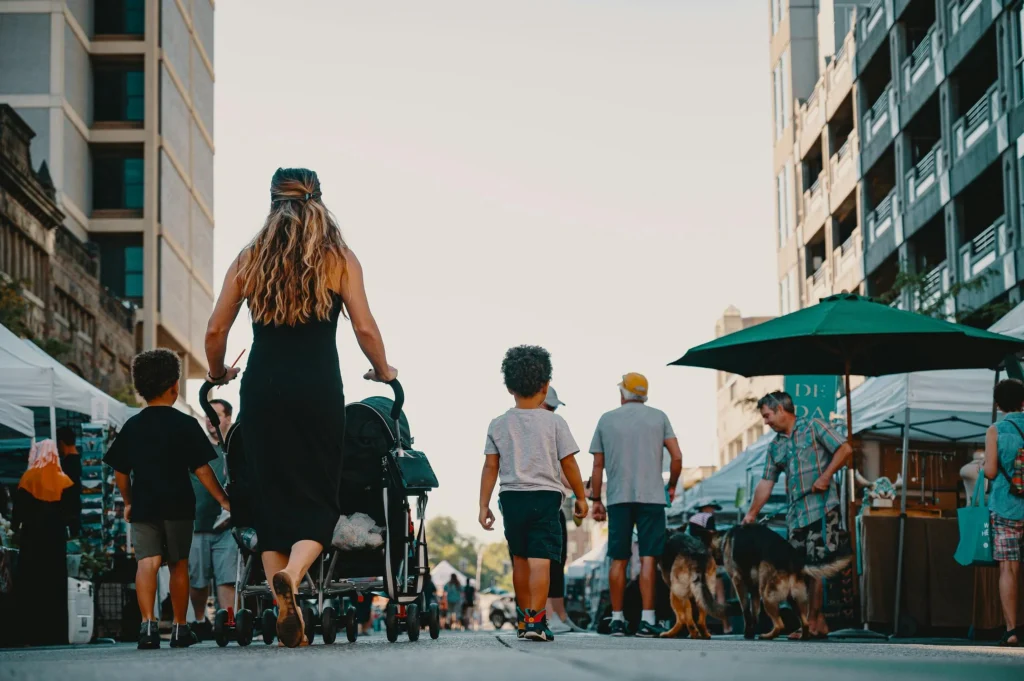 An image showing a woman on a walkable vendor-filled street with a stroller and two small children next to her. 