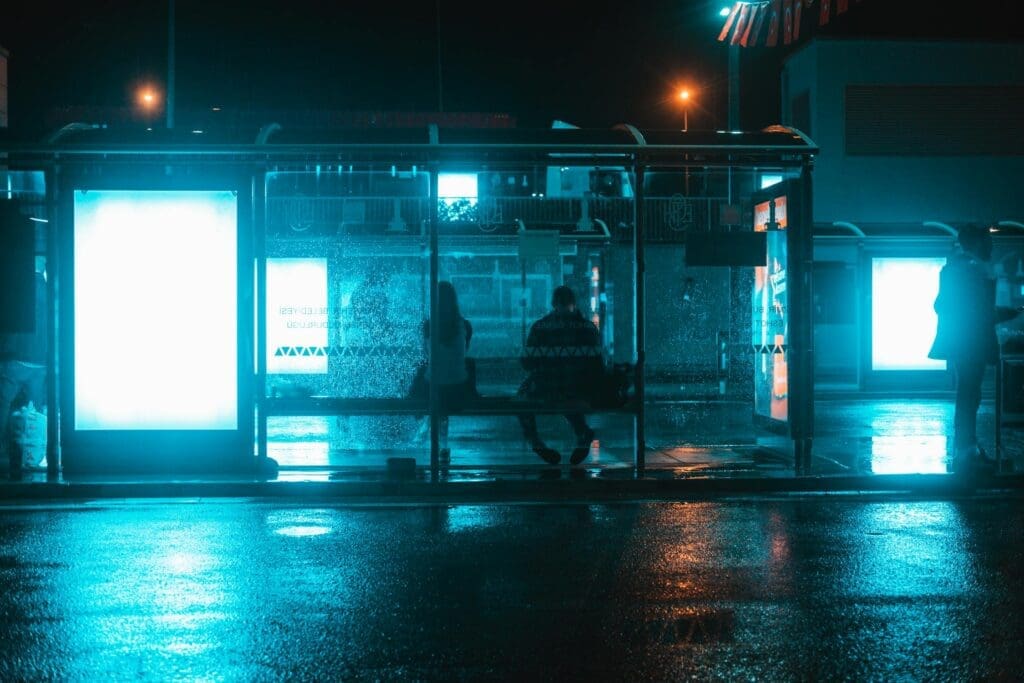 Una parada de autobús con luz azul oscura y dos personas sentadas en un banco, esperando.