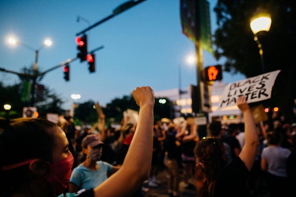 Una protesta llena de gente. En el centro se ve a una persona con el puño en alto y a otra con un letrero de Black Lives Matter.